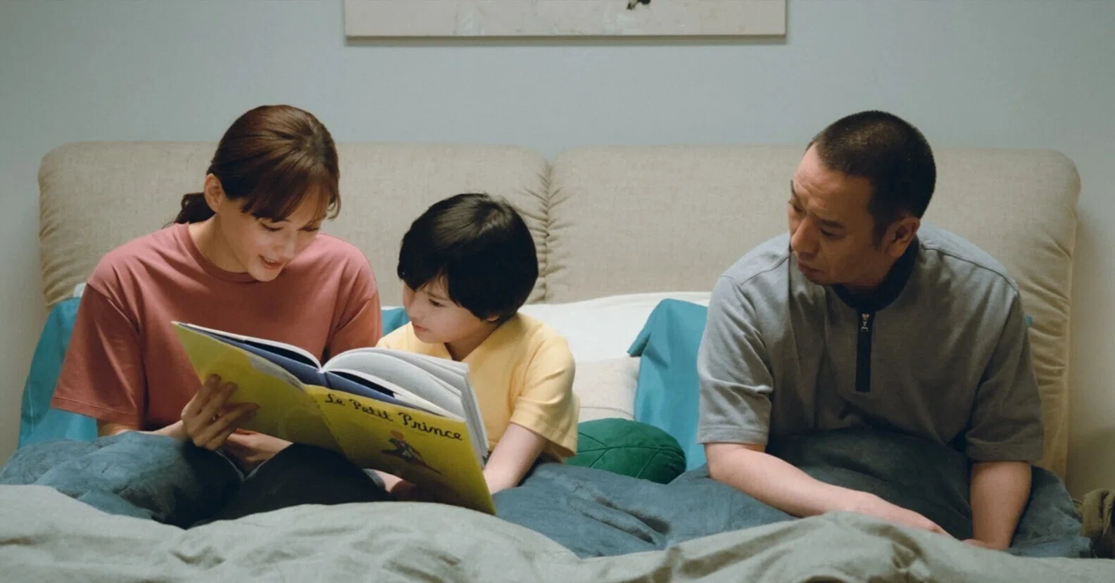 Mom reading "The Little Prince" to boy in bed while Father watches from other side.